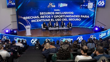 Cinco personas sentadas en un escenario frente a una pantalla gigante azul con texto sobre seguros, ante una audiencia en un auditorio