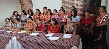 Grupo de mujeres con vestimenta tradicional colorida, sentadas y de pie en una reunión. Mesa con mantel tejido. Una mujer habla por micrófono