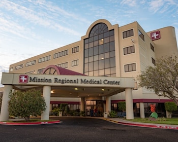 Fachada de un edificio de varias plantas con el cartel 'Mission Regional Medical Center', cruces rojas, ventanas y vegetación