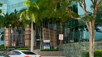 Detalhamento em vidro e pedra da nova Prefeitura de Fort Lauderdale, com palmeiras e árvores. Uma parte do carro branco pode ser vista em primeiro plano