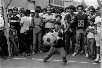 Un niño patea un balón de fútbol en un juego informal, con una multitud de adultos y niños observando detrás. Imagen en blanco y negro