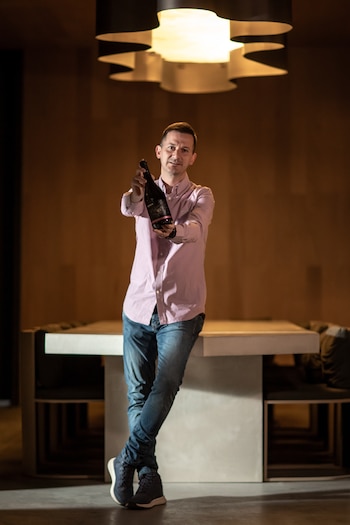 Hombre de pie, camisa rosa y jeans, extiende una botella de vino oscuro hacia adelante. Detrás, una mesa y una lámpara de diseño en un interior con luz cálida