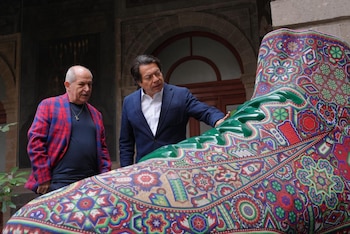 Dos hombres, uno con traje azul y otro con saco a cuadros, observan una gran escultura en forma de bota decorada con patrones coloridos wixárika