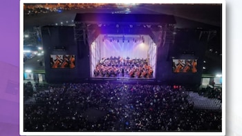 Escenario de concierto al aire libre con una orquesta sinfónica, director, pantallas laterales y un gran público. Luces iluminan el escenario