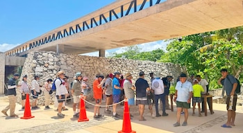 Grupo de turistas haciendo fila al aire libre en una zona arqueológica, delimitada por conos naranjas y cuerdas, con un muro de piedra, vegetación y una estructura de concreto al fondo