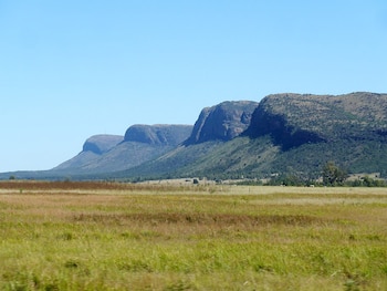 Cadena de montañas con laderas escarpadas y vegetación, situada tras una extensa llanura de pastizales secos bajo un cielo azul despejado