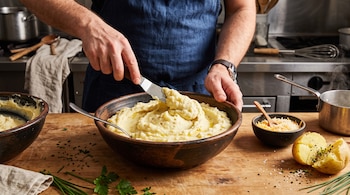 Manos de un cocinero mezclan puré en un cuenco con una espátula, junto a una papa cocida, queso rallado y hierbas sobre una mesa de madera.