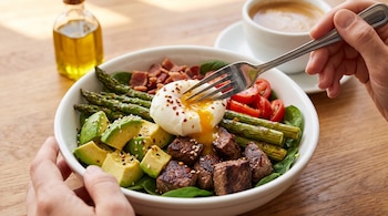 Persona sujetando un plato blanco con tenedor sobre un huevo poché, aguacate, carne, espárragos, tocino, tomates, hojas verdes, sésamo, ají, aceite y café