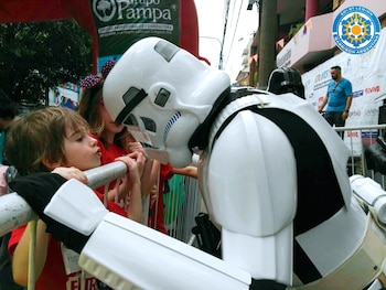Un niño con cabello castaño claro se apoya en una valla metálica gris y mira con curiosidad a un cosplayer de Stormtrooper, quien lleva casco y armadura blanca