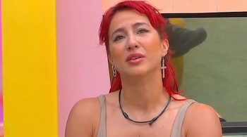Primer plano de una mujer de cabello rojo, camiseta clara, collar negro y pendiente en forma de cruz sobre un fondo rosa y amarillo.