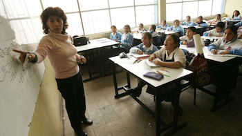 Una profesora señala una pizarra con ecuaciones frente a un grupo de alumnas en un aula con ventanas amplias