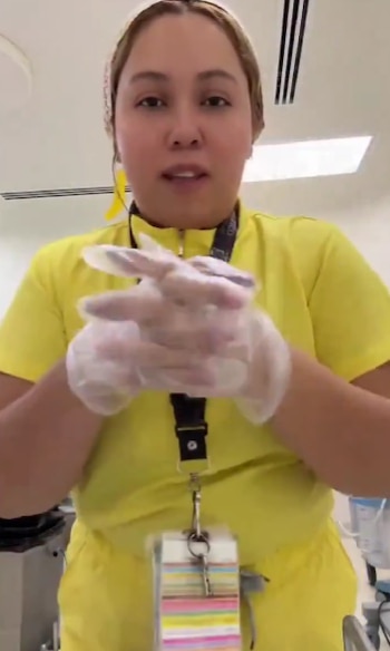 Una mujer con uniforme médico amarillo junta sus manos cubiertas con guantes transparentes y holgados en una habitación blanca
