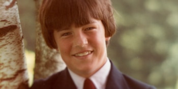 Primer plano de un joven sonriente con cabello castaño, saco oscuro, camisa blanca y corbata junto al tronco de un abedul