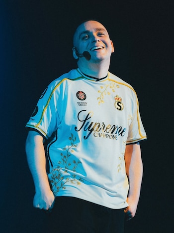 Lucho Miranda, un hombre calvo con micrófono de diadema, sonríe ampliamente. Viste una camiseta blanca con letras y patrones dorados, manos en bolsillos, sobre fondo oscuro