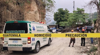 Vista trasera de una ambulancia de Sanarate y cinta amarilla con texto "HOMICIDIO PRECAUCIÓN" en un camino de tierra, con personas y vehículos al fondo.