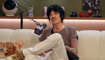 Tommy Lee sentado en un sofá claro, con camiseta gris y pantalones claros, cubierto de tatuajes. Un micrófono frente a él, un perro pequeño en el sofá