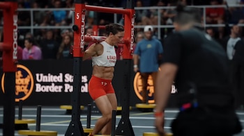 Tia-Clair Toomey, atleta, realiza una sentadilla con barra acolchada sobre sus hombros en un evento de CrossFit, vistiendo top blanco y shorts rojos