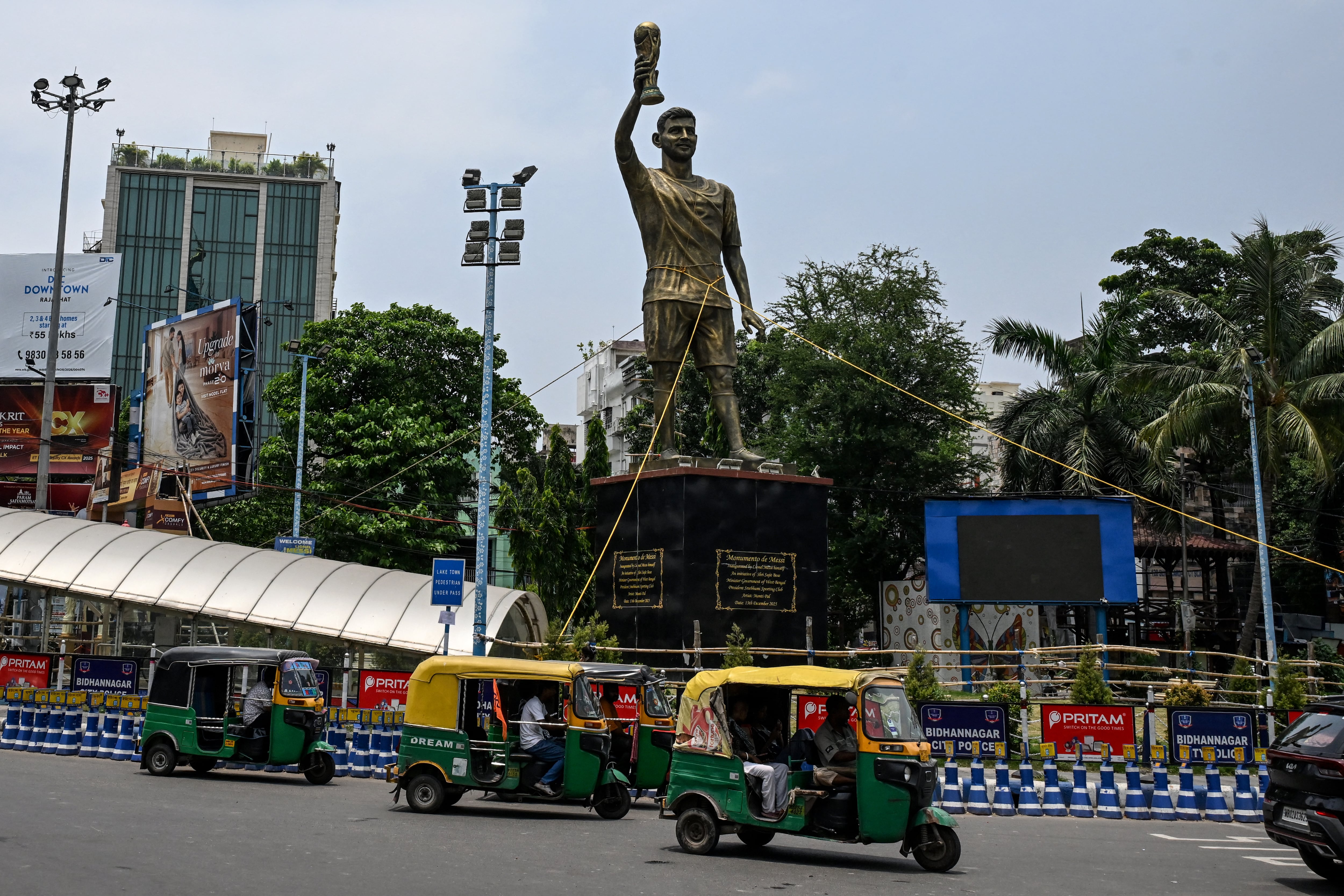 Los rickshaws pasan junto a una estatua del futbolista argentino Lionel Messi que ha sido atada con una cuerda para protegerla de la lluvia y las tormentas, antes de su reinstalación en un lugar más seguro, en Calcuta el 27 de mayo de 2026 (Foto de Dibyangshu SARKAR / AFP)