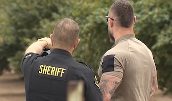 Dos hombres vistos de espalda en una zona arbolada, uno con uniforme negro que dice SHERIFF y otro con camiseta beige y tatuajes