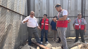 Varias personas, incluyendo hombres con chalecos de emergencia, inspeccionan una zona dañada con una valla de alambre caída y escombros