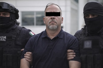 Un hombre central con barba y los ojos cubiertos por una barra negra es escoltado por dos policías uniformados con equipo táctico.