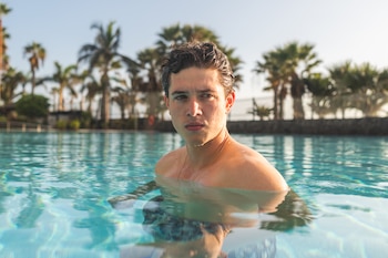 Un joven con cabello oscuro mojado y gotas de agua en la piel emerge de una piscina azul. El fondo borroso muestra palmeras bajo un cielo claro