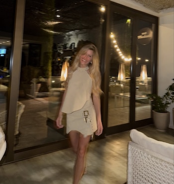 Una mujer rubia de cabello largo sonríe mientras camina en un espacio nocturno iluminado por luces cálidas, vistiendo un conjunto beige de dos piezas