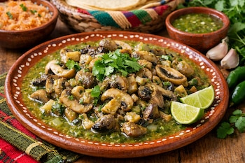 Plato de barro con setas en salsa verde, cilantro y rodajas de lima. Se observan cuencos de arroz, tortillas, salsa verde, ajos y chiles.