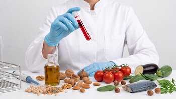 Una persona con bata sostiene un tubo con líquido rojo ante una mesa con aceite, frutos secos, tomates, pescado, legumbres y hortalizas