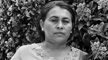 Retrato de primer plano medio de una mujer vestida con una blusa de encaje frente a un arbusto florecido en blanco y negro.