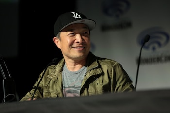Primer plano de un hombre con gorra negra con logo blanco 'LA', camiseta gris y chaqueta verde oliva, sonriendo frente a un micrófono y un fondo oscuro