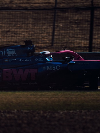 Un coche de Fórmula 1 azul oscuro y rosa con varios logotipos de patrocinadores, un conductor en el habitáculo, en una pista de carreras