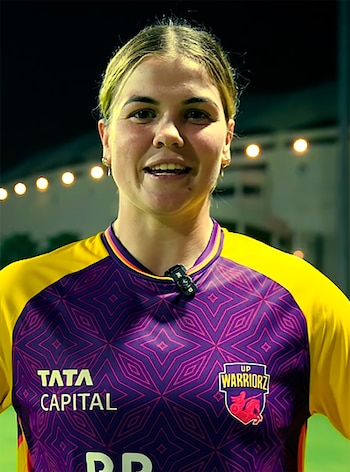 Georgia Voll sonríe con uniforme deportivo amarillo y morado, logos de Tata Capital y UP Warriorz, un micrófono en el cuello y fondo oscuro