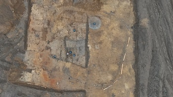 Vista superior de un terreno excavado con cubos azules, hendiduras rectangulares, estacas de madera en arco y una pala