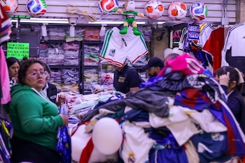 Interior de un local comercial con estantes llenos de ropa, balones de fútbol colgando y un letrero de "Abierto". Varias personas examinan pilas de prendas deportivas en mesas