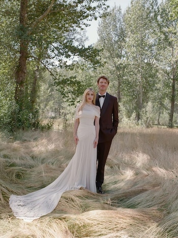 Mujer rubia con vestido de novia blanco largo y hombre con traje oscuro y pajarita posan de pie en un campo de hierba seca con árboles al fondo