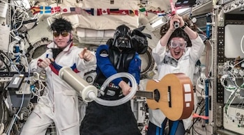Tres personas en una estación espacial con peluca, gafas, una máscara, una guitarra acústica, un tubo flexible y banderas colgadas