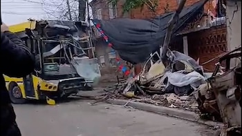 Un colectivo amarillo con la parte delantera destruida junto a los restos de un vehículo aplastado frente a una vivienda
