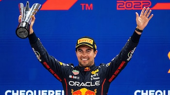 Sergio Pérez sonríe con una gorra y un traje de carreras de Red Bull mientras sostiene un trofeo plateado y saluda con la mano alzada.