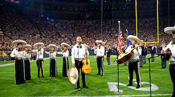 Integrantes de un mariachi con trajes tradicionales, sombreros e instrumentos musicales sobre el césped de un estadio de fútbol americano.
