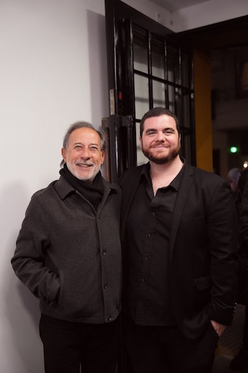 Dos hombres, Guillermo Francella con abrigo gris y bufanda negra junto a Gastón Cocchiarale con traje negro, sonríen frente a una puerta oscura