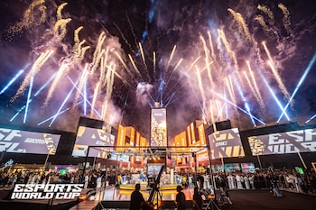 Un escenario al aire libre con pantallas gigantes, luces de colores, fuegos artificiales en el cielo y personas reunidas por la noche
