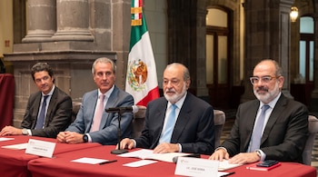 Cuatro hombres con traje y corbata están sentados frente a una mesa con mantel rojo, micrófonos, identificadores y la bandera de México.