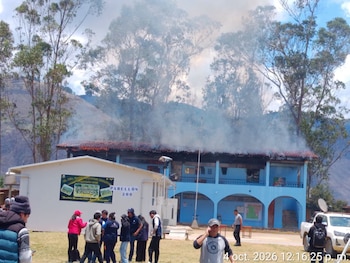 Varias personas permanecen en un terreno de hierba ante un edificio azul con humo saliendo de su techo y montañas en el fondo