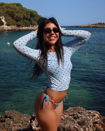 Mujer de cabello oscuro con gafas de sol, traje de baño celeste de lunares y braga con lazos naranjas, posa sobre rocas frente al mar y vegetación en un día soleado