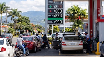 Una gasolinera con un letrero digital de precios, varios vehículos haciendo fila y personal de servicio en un día soleado.