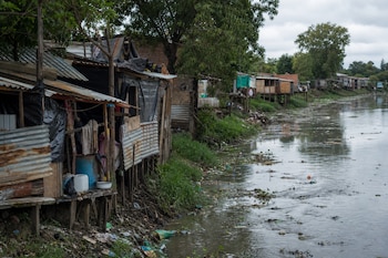 Viviendas de chapa y madera construidas sobre la orilla de un río con vegetación y basura acumulada en el agua.