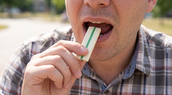 Primer plano de un hombre que introduce un chicle rectangular blanco con una raya verde en su boca, sus dientes y parte de la lengua visibles.