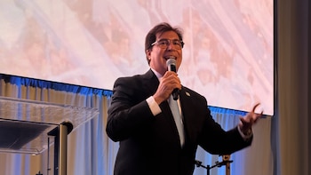 Lisandro Carret, un hombre sonriente con gafas, traje oscuro y corbata clara, habla en un micrófono plateado, con una pantalla luminosa detrás
