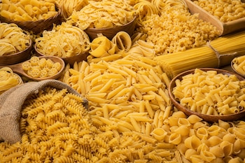 Variedad de pasta seca como espaguetis, espirales, lazos y plumas dispuestos en cuencos de madera, sacos y sobre una mesa.